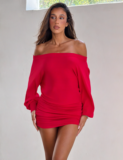 A woman with long brown hair stands indoors in the TIGERMIST JANINE MINI DRESS - RED, an off-shoulder, long-sleeve knit dress with a fitted silhouette. She gazes slightly to the side as light streams in from a window above.
