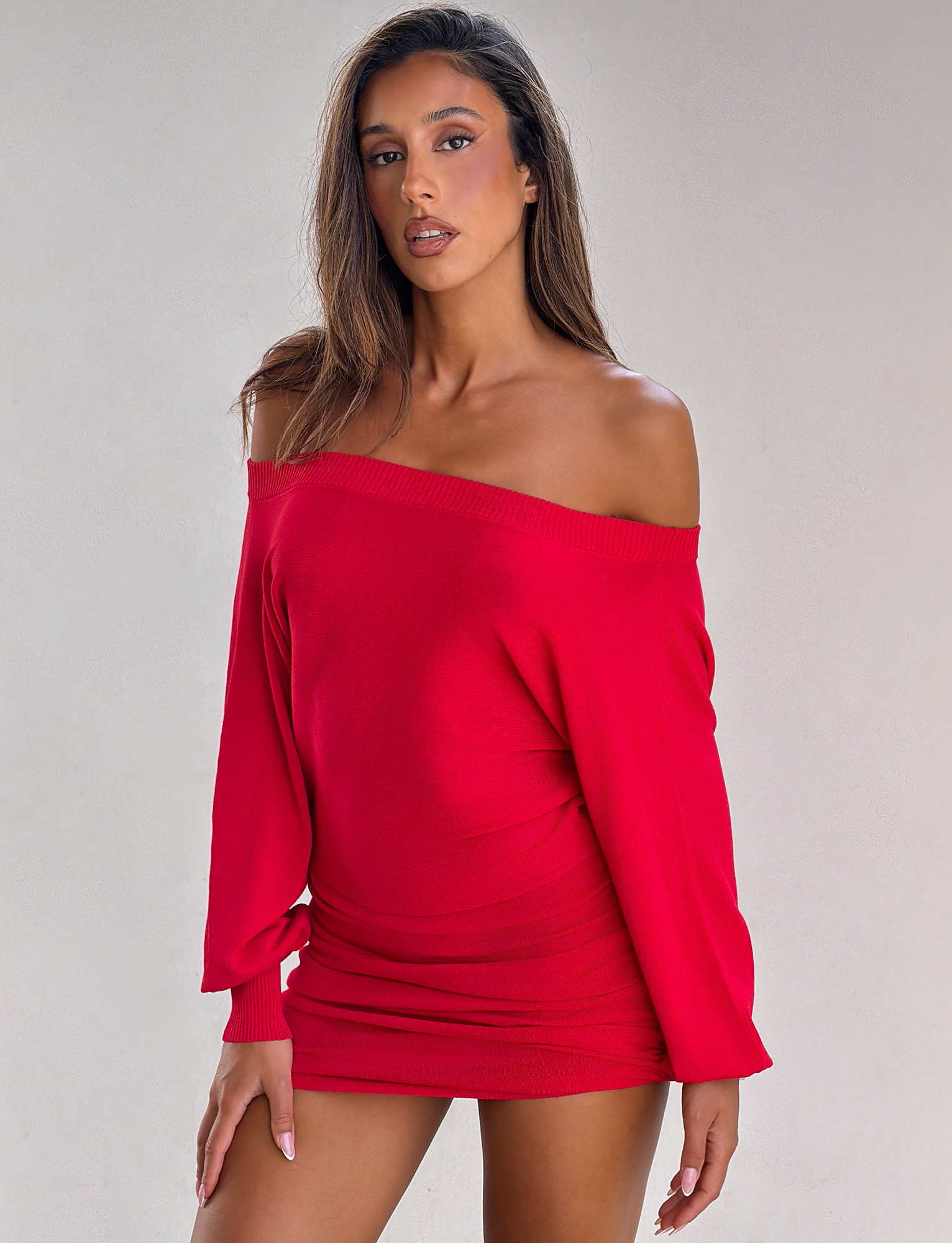 A woman models the TIGERMIST JANINE MINI DRESS in red, featuring off-shoulder long sleeves, standing confidently against a plain light background.
