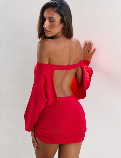 A woman poses against a white wall in the TIGERMIST JANINE MINI DRESS - RED, showcasing its off-the-shoulder neckline, long sleeves, ruched bodycon mini skirt, and open back as she turns slightly away.