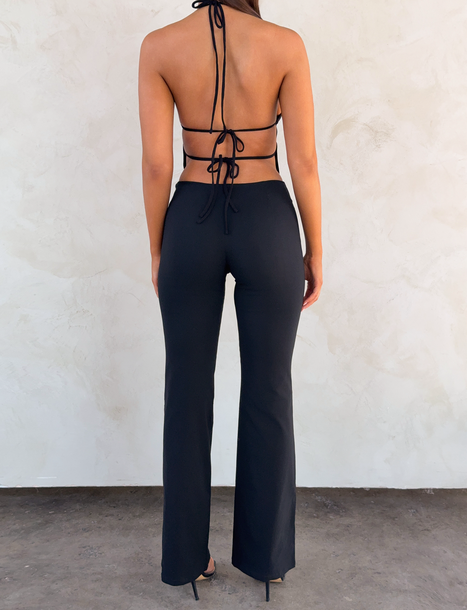 A woman faces away from the camera, wearing TIGERMIST's MARREN PANT in black and a black halter top with an open, tie-back design, standing on a dark floor against a textured light wall.