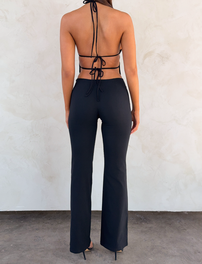 A woman faces away from the camera, wearing TIGERMIST's MARREN PANT in black and a black halter top with an open, tie-back design, standing on a dark floor against a textured light wall.
