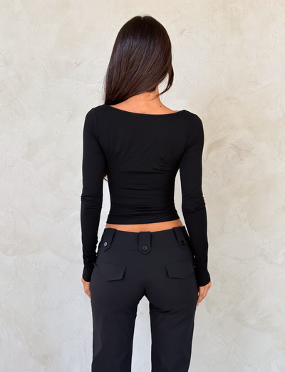 A woman with long dark hair, seen from behind, wears the TIGERMIST DAMELA LONG SLEEVE TOP in black with an adjustable neckline and fitted black pants featuring buttoned pockets, standing against a light textured wall.