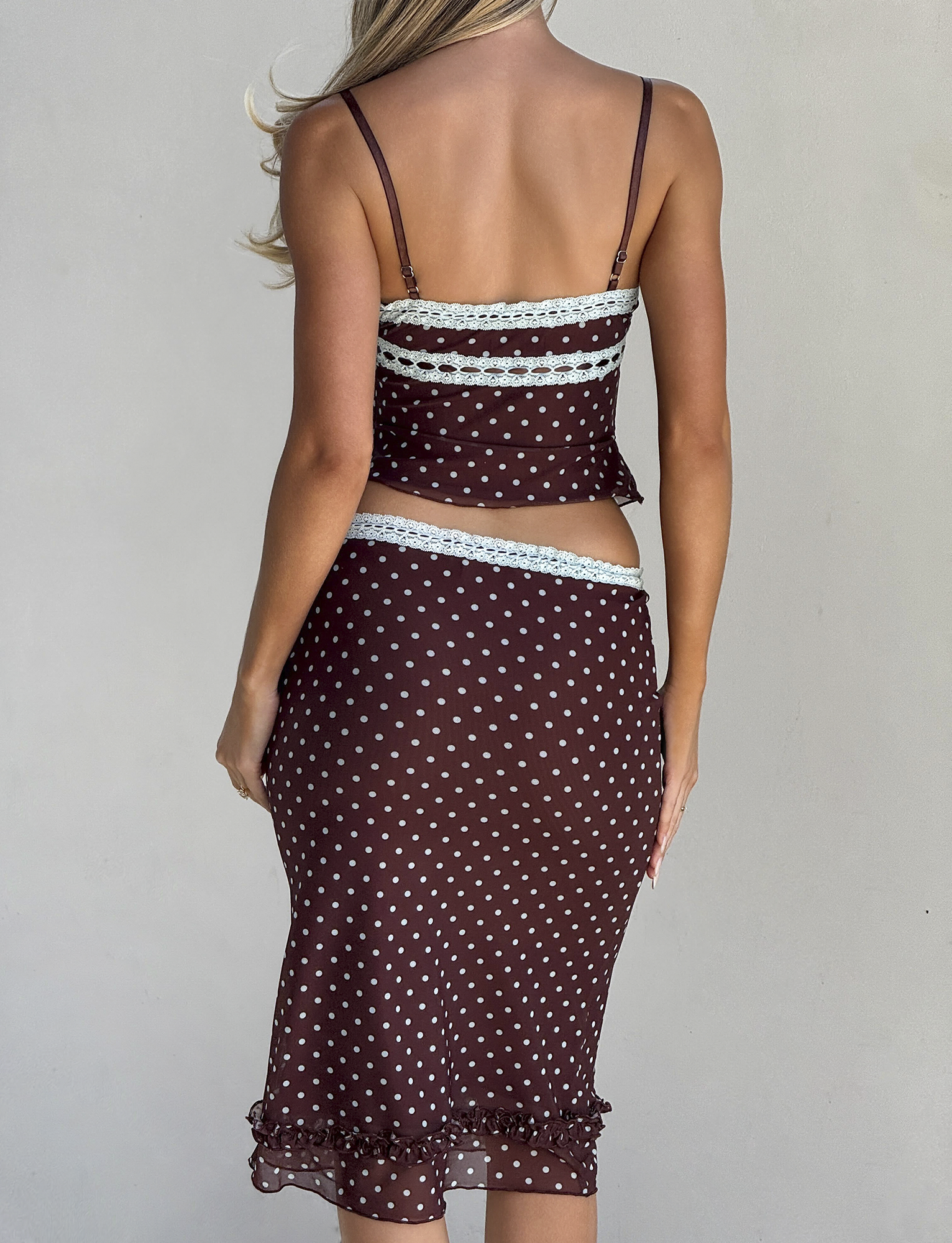 A woman with long, wavy hair is seen from behind wearing the TIGERMIST AMARA TOP - POLKA DOT, a brown two-piece with light blue lace trim and sheer mesh cami, paired with a matching midi skirt.