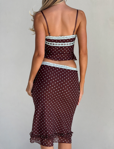 A woman with long, wavy hair is seen from behind wearing the TIGERMIST AMARA TOP - POLKA DOT, a brown two-piece with light blue lace trim and sheer mesh cami, paired with a matching midi skirt.