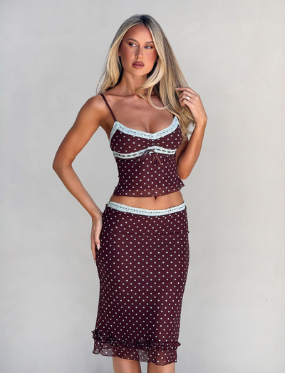 A woman with long blonde hair poses against a light background, wearing the TIGERMIST AMARA MIDI SKIRT - POLKA DOT and matching spaghetti strap top, both brown with white dots and light blue lace trim.