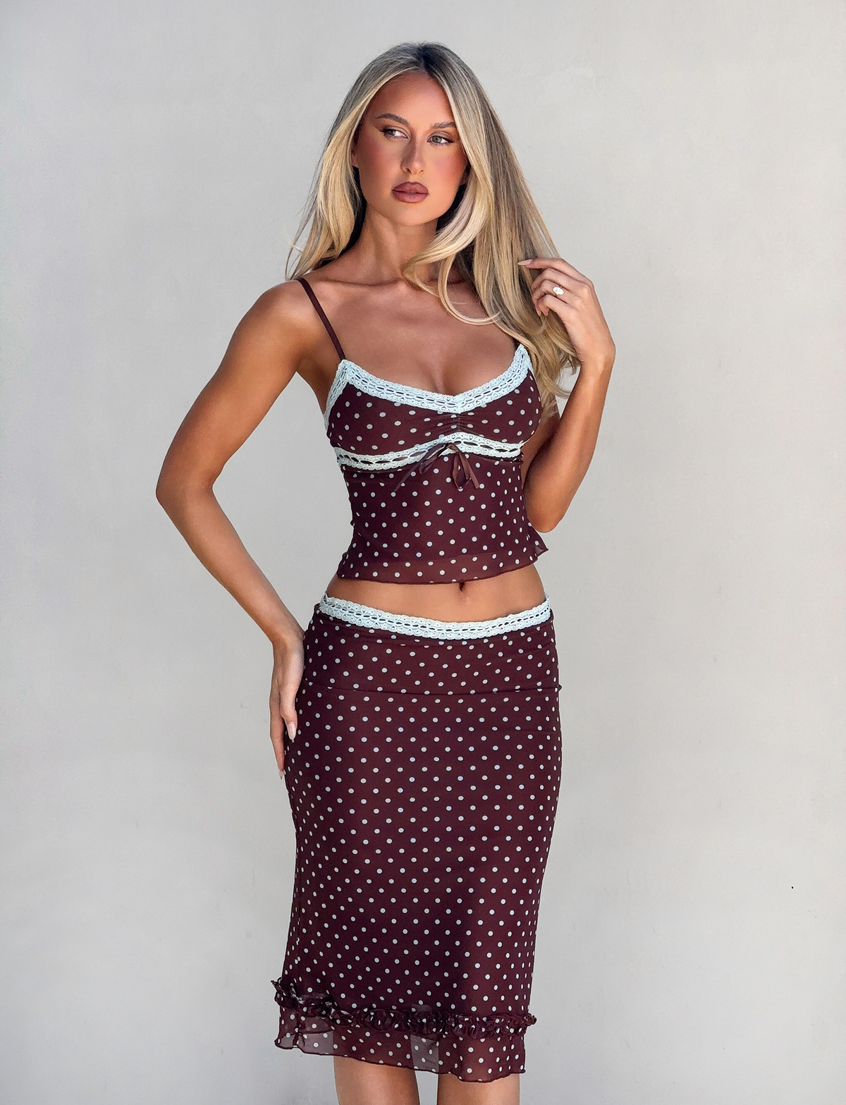 A woman with long blonde hair wears the TIGERMIST AMARA TOP - POLKA DOT, a fitted brown camisole with pale blue lace trim, paired with a matching skirt, standing against a plain light gray background.