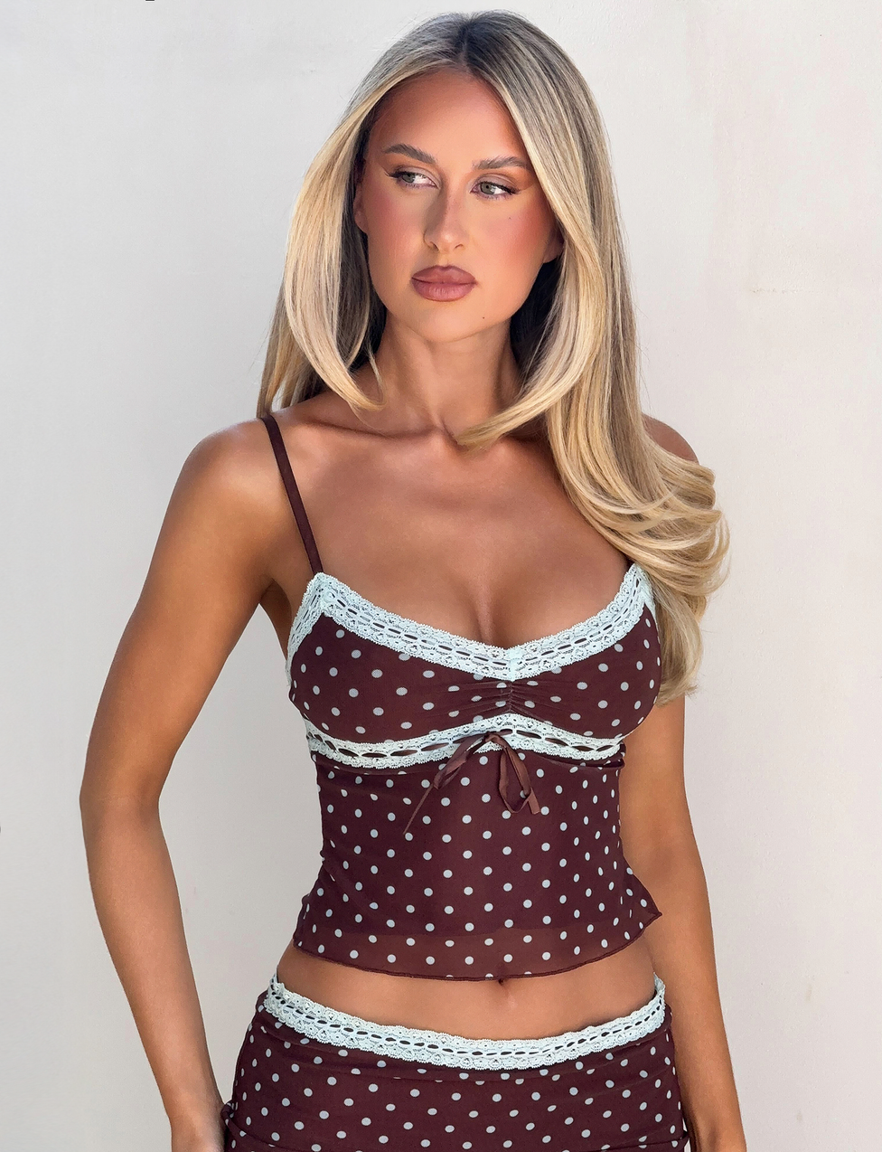 A woman with long blonde hair models the AMARA TOP - POLKA DOT by TIGERMIST, featuring delicate white lace trim. She stands against a plain light background, gazing slightly left with a neutral expression.