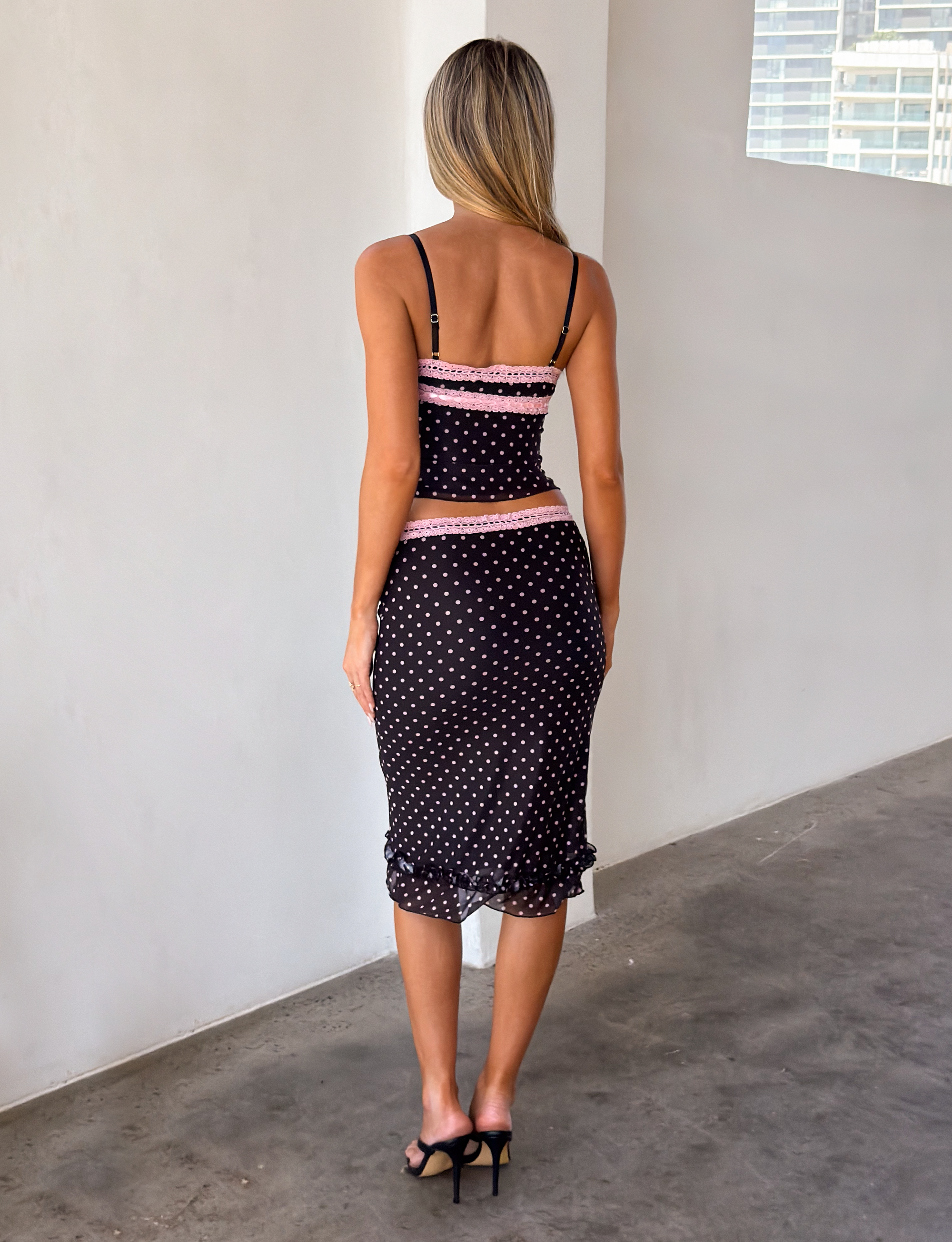 A woman with long, straight blonde hair stands indoors on a smooth concrete floor with white walls, wearing the TIGERMIST AMARA TOP in black and black high-heeled sandals. She is facing away from the camera.