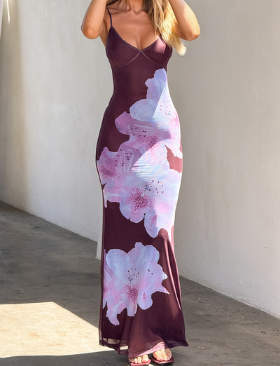 A woman stands against a light wall in the TIGERMIST ORIELLE MAXI DRESS - PLUM ORCHID, a sleeveless maxi featuring large pastel pink and white floral placement prints, paired with open-toe sandals. Her face is partially obscured.