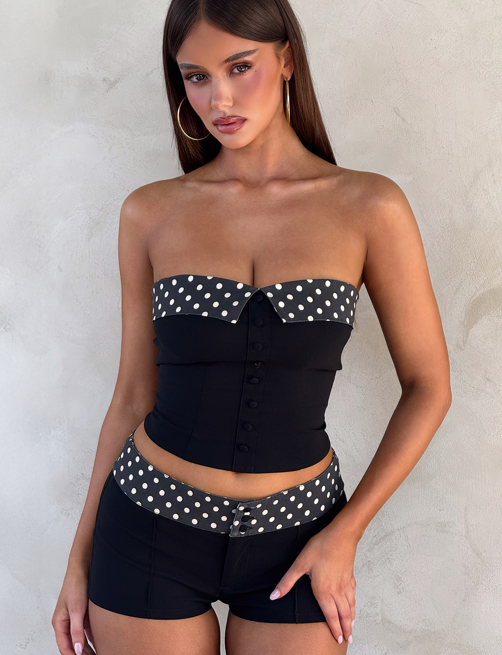 A woman with long brown hair models the TIGERMIST KITTIE 2.0 BUSTIER in black, paired with matching black shorts featuring black and white polka dot trim, made from elegant suiting fabric, against a light textured background.