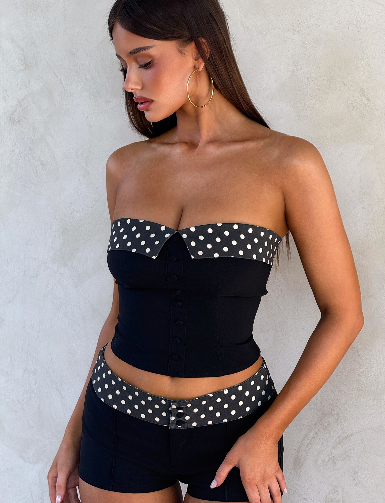 A woman with long brown hair and large hoop earrings stands against a light textured wall wearing the TIGERMIST KITTIE 2.0 BUSTIER - BLACK, featuring a strapless design with a polka dot band, paired with matching high-waisted shorts.
