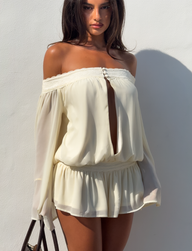 A woman poses against a white wall wearing the TIGERMIST TESSIE PLAYSUIT in ivory, featuring an off-shoulder neckline, long sleeves, front cutout, and ruffled hem. She carries a black bag as sunlight casts shadows behind her.
