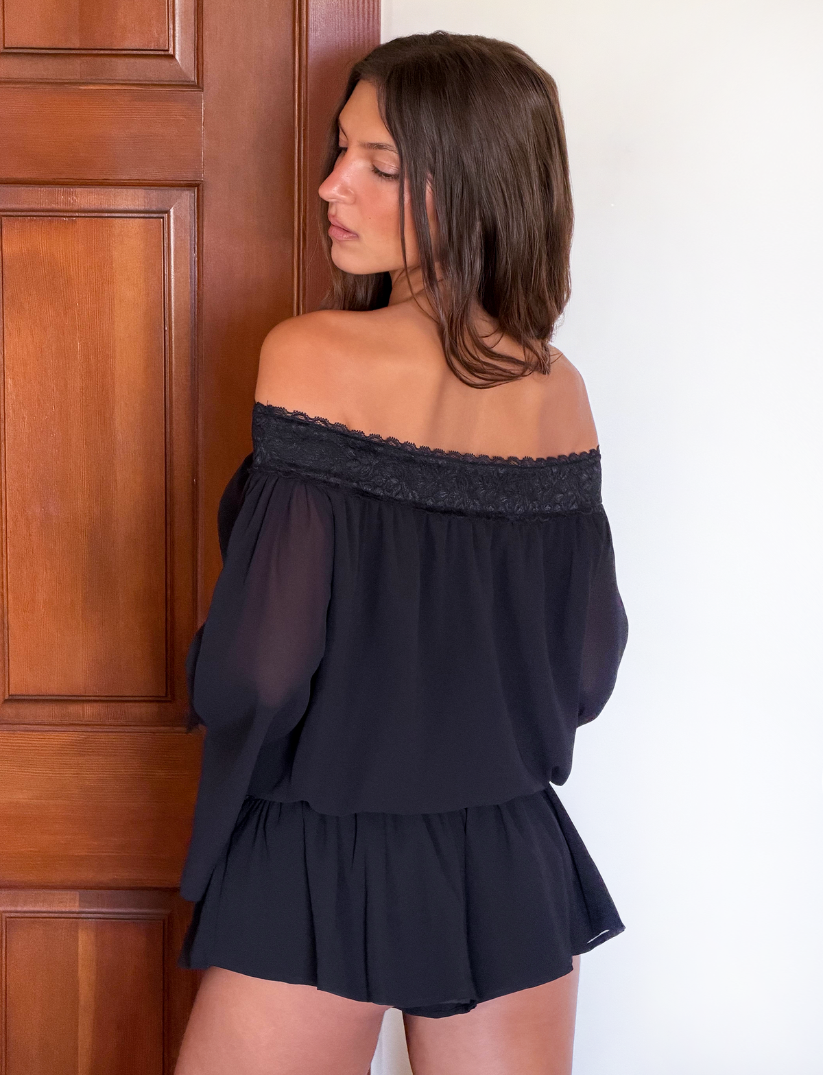 A woman with long brown hair stands indoors by a wooden door, wearing the TIGERMIST TESSIE PLAYSUIT in black with lace trim, and looks over her shoulder.