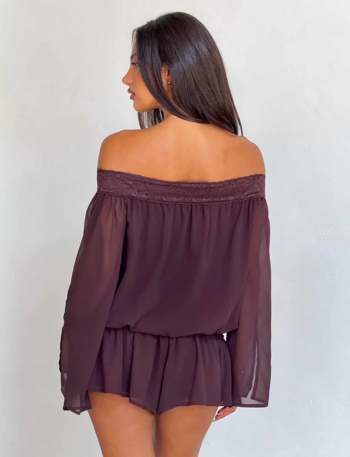 A woman with long dark hair faces away, wearing the TIGERMIST TESSIE PLAYSUIT in brown—an off-shoulder design with sheer fabric, lace details, and ruffled mini shorts—set against a light gray background.
