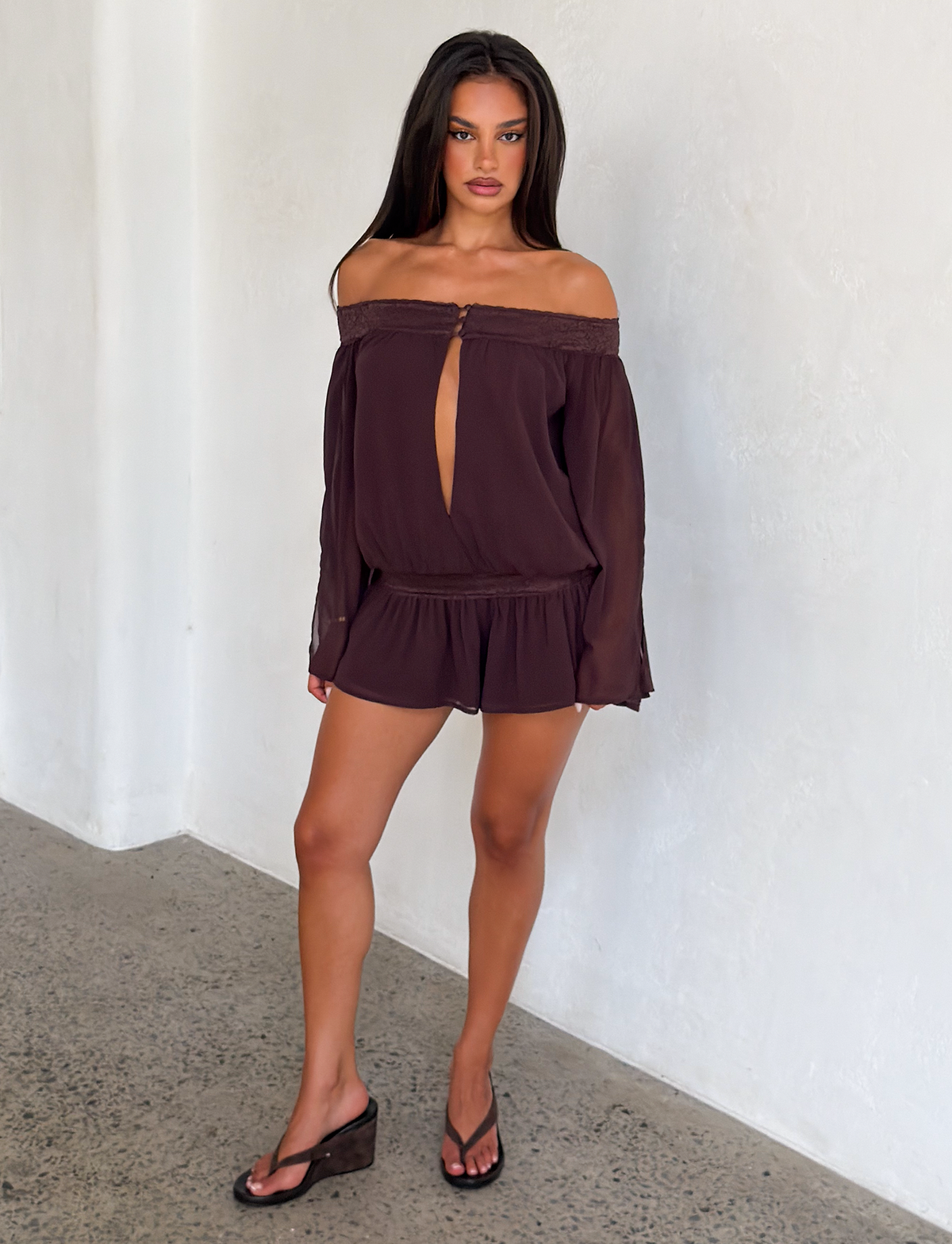 A woman stands indoors against a white wall, wearing the TIGERMIST TESSIE PLAYSUIT in brown—an off-shoulder style with long sleeves, front keyhole cutout, and ruffled mini shorts. She pairs it with wedge sandals and flip-flops.
