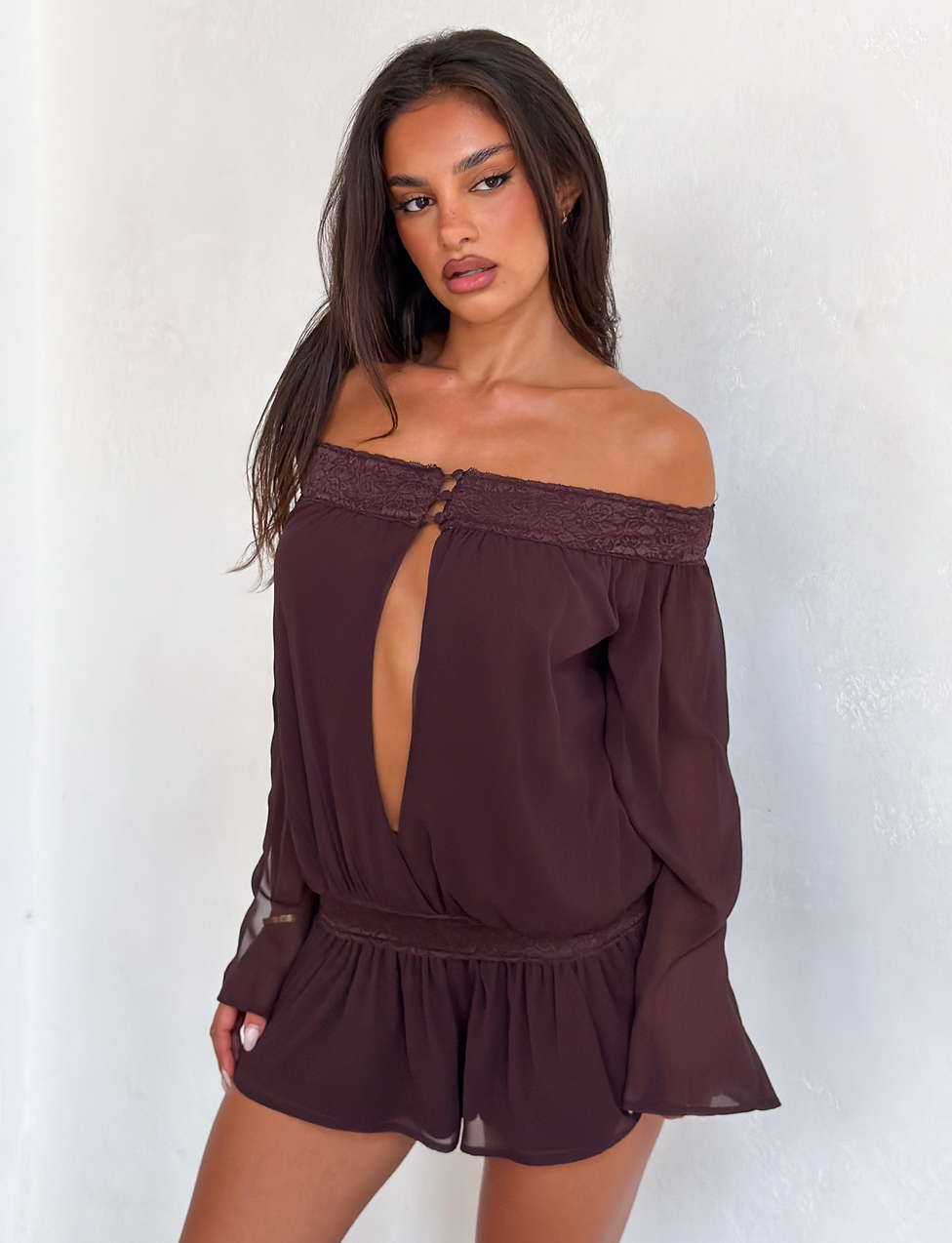 A woman with long brown hair models the TIGERMIST TESSIE PLAYSUIT in brown, featuring ruffled mini shorts and a front cutout, set against a plain white background.
