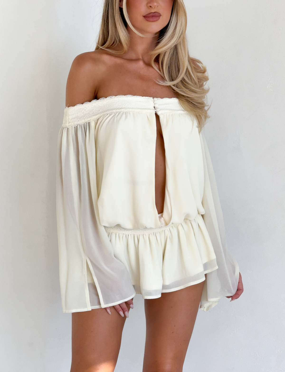 A woman models the TIGERMIST TESSIE PLAYSUIT in ivory, featuring an off-shoulder neckline, long flowy sleeves, ruffled micro mini shorts, and a front keyhole cutout, standing against a plain white background.
