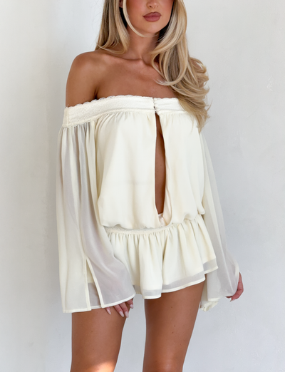 A woman models the TIGERMIST TESSIE PLAYSUIT in ivory, featuring an off-shoulder neckline, long flowy sleeves, ruffled micro mini shorts, and a front keyhole cutout, standing against a plain white background.