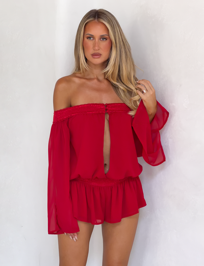 A woman with long blonde hair wears the TIGERMIST TESSIE PLAYSUIT in red, featuring long sleeves, ruffled mini shorts, and a keyhole front, as she stands against a white background looking at the camera.