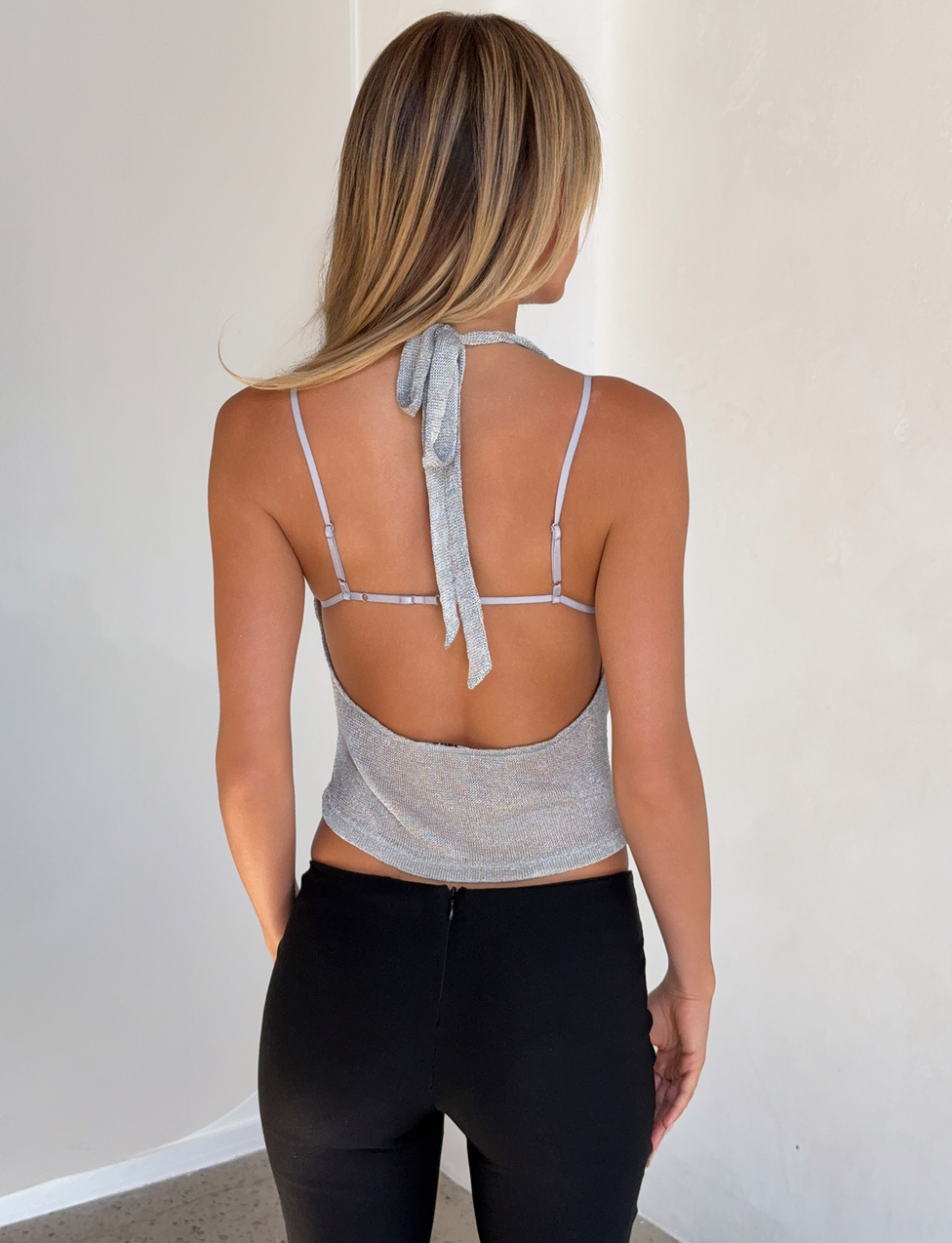 A woman with long blonde hair faces away indoors, wearing the TIGERMIST MAKENA TWO-PIECE TOP 2.0 in silver with an open back and black pants, standing near a plain light-colored wall.