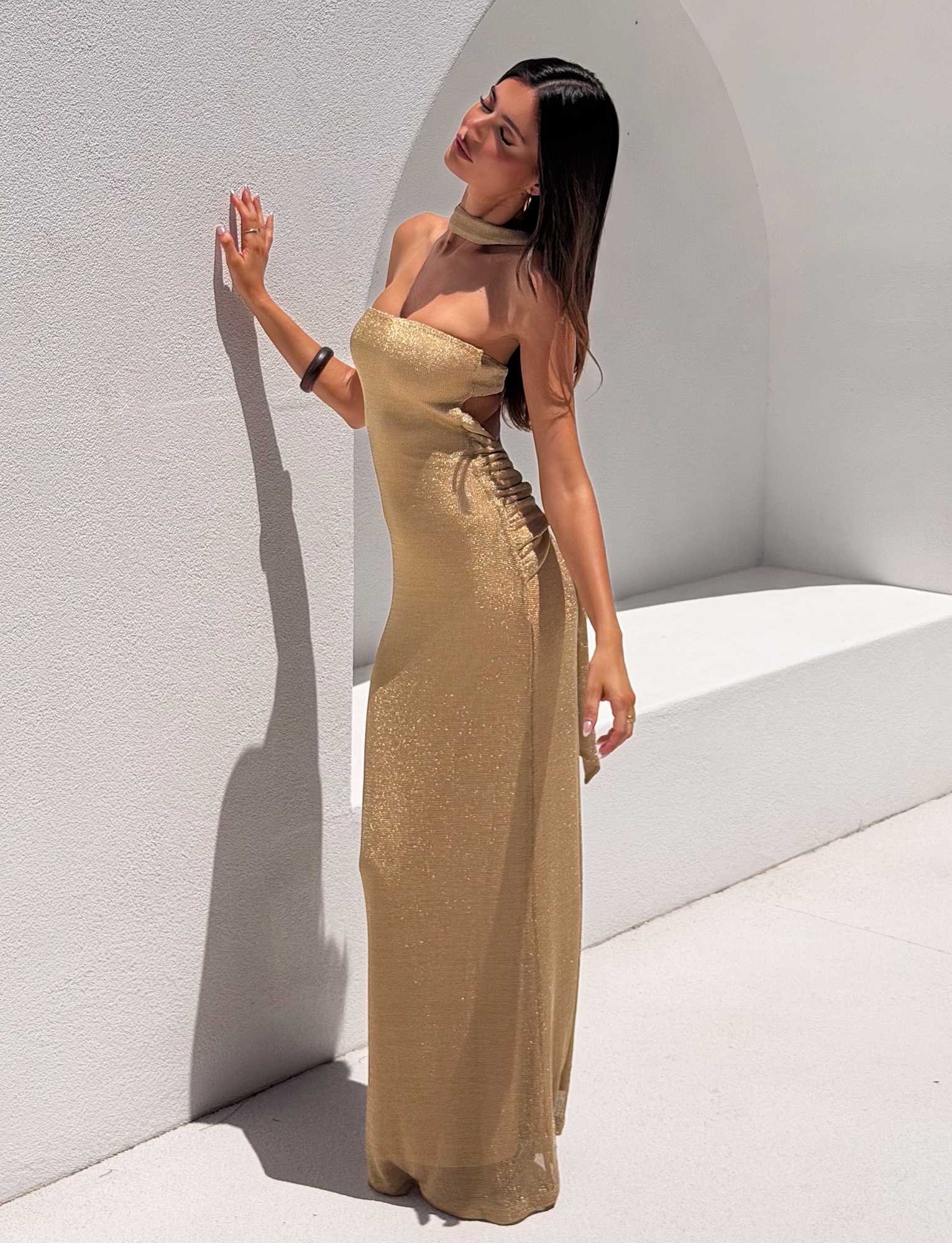 A woman models the DANIELA SCARF DRESS 2.0 - GOLD by TIGERMIST, posing elegantly against a white curved wall with one hand resting on it, head tilted back and eyes closed in bright sunlight.
