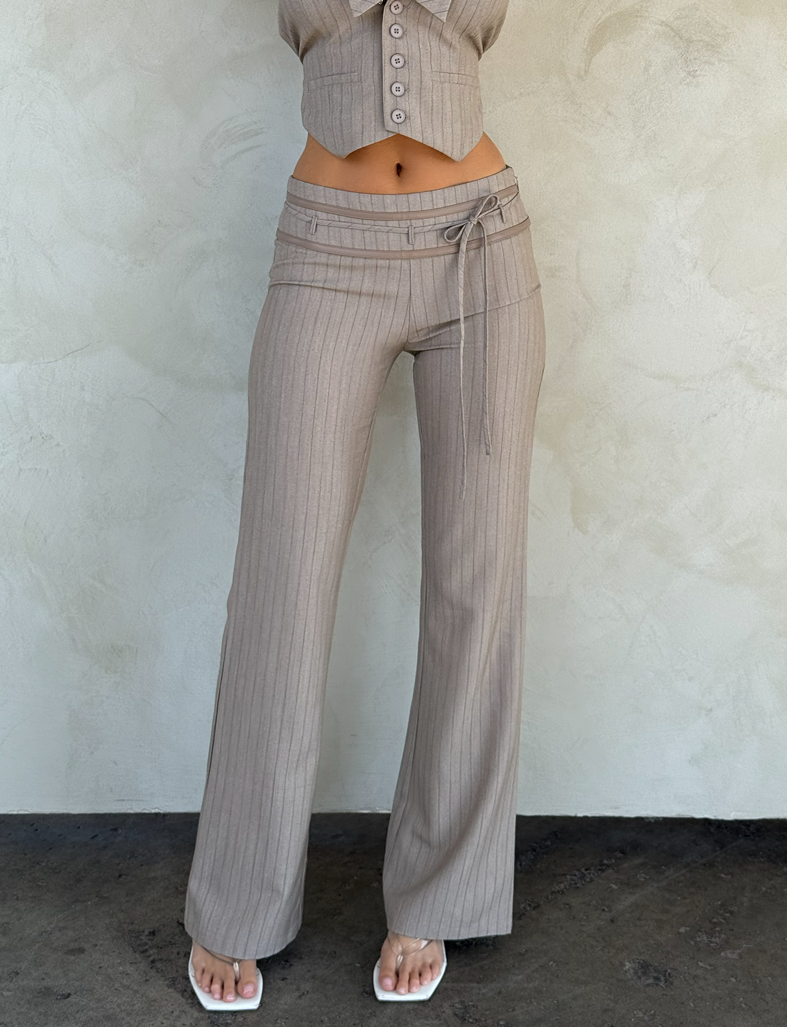 Someone wears the TIGERMIST FEDRA PANT - TAN PINSTRIPE with a tie belt and a matching cropped top, posed against a light textured wall. Their head is cropped out of the image.