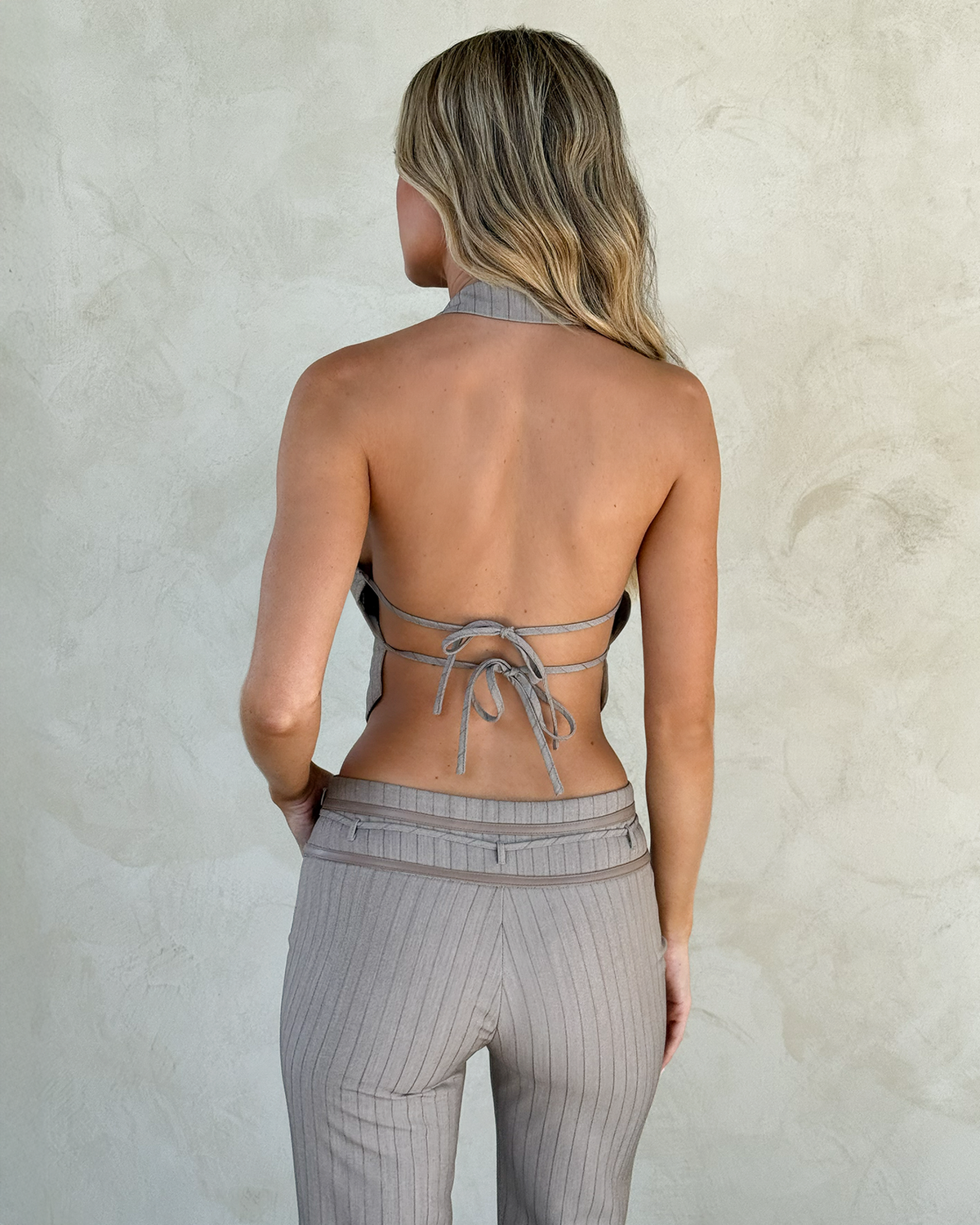 A woman with long, wavy blonde hair is seen from the back wearing the TIGERMIST FEDRA VEST - TAN PINSTRIPE halter vest, tied at the back, with matching high-waisted pants against a light textured wall.