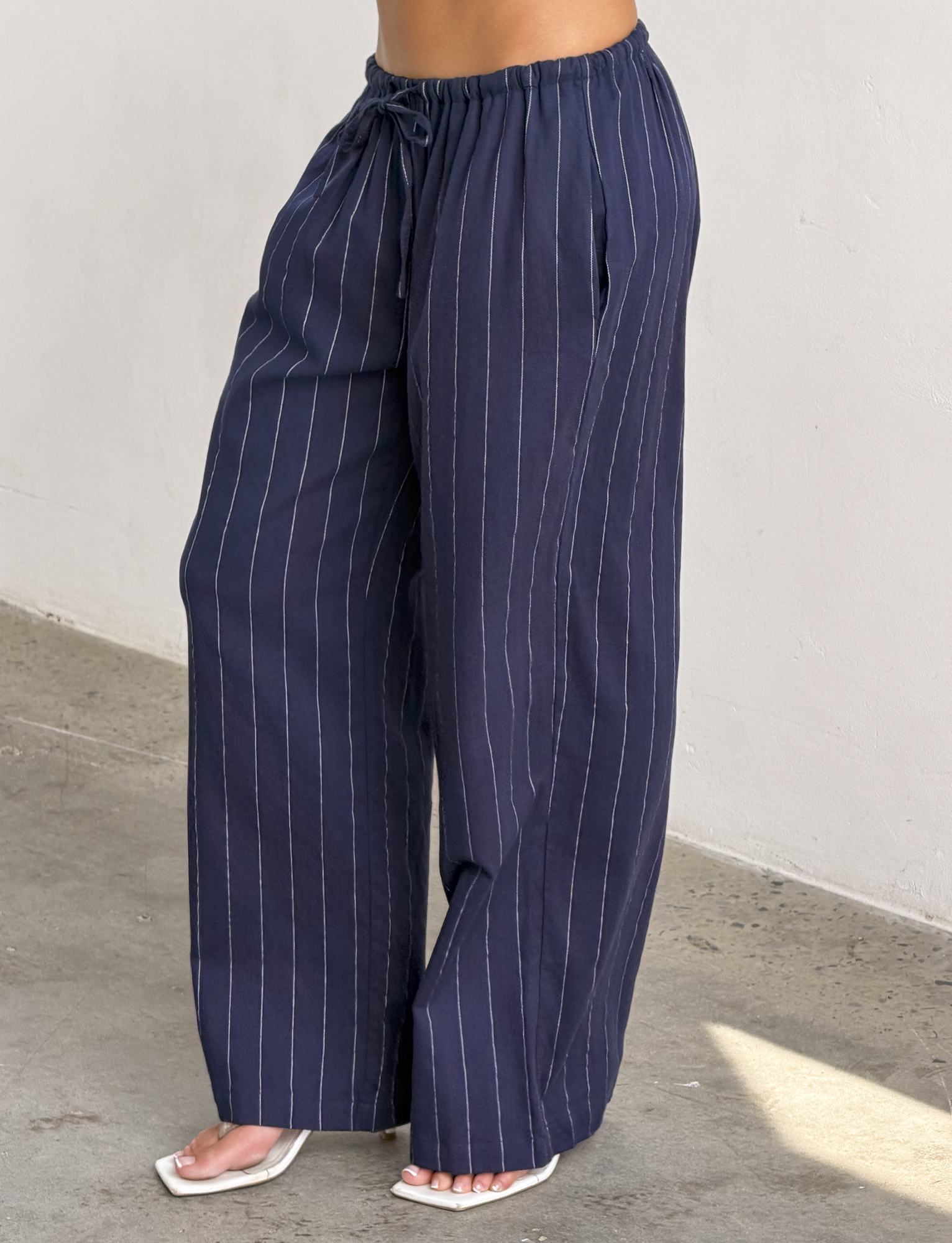 A person wears the TIGERMIST LAINEY PANT in navy pinstripe with an elasticated waist and white open-toe heels, standing on a concrete floor against a plain light wall.