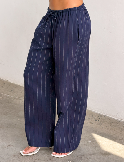 A person wears the TIGERMIST LAINEY PANT in navy pinstripe with an elasticated waist and white open-toe heels, standing on a concrete floor against a plain light wall.