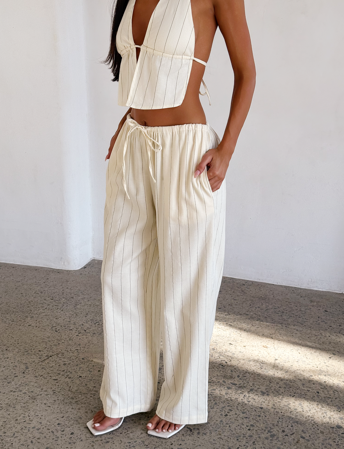A woman stands indoors on a speckled floor, wearing the TIGERMIST LAINEY PANT - BEIGE STRIPE with a matching light-colored halter top, her hand in her pocket.