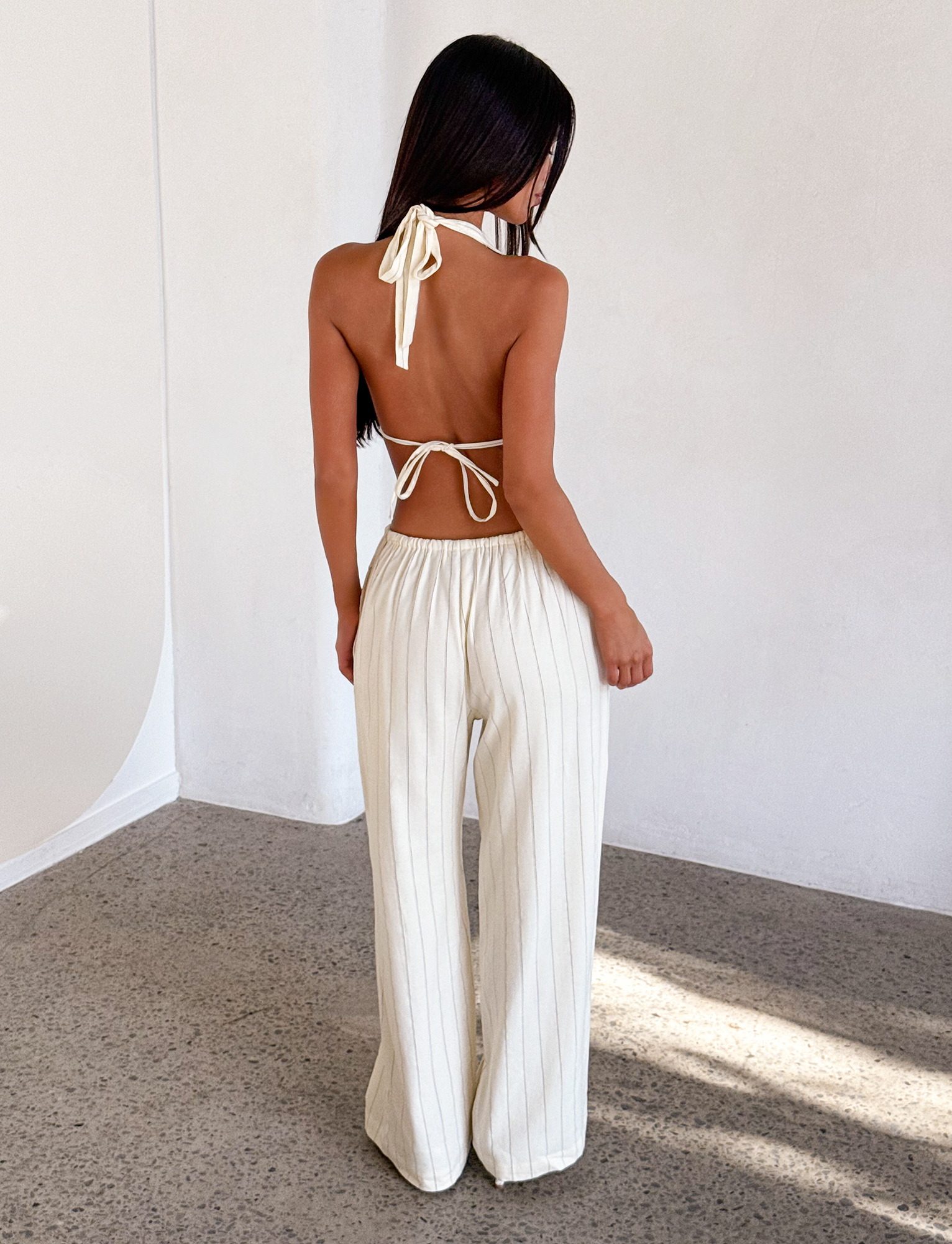 A woman with long dark hair stands indoors in a minimalist space, wearing a backless halter top and the TIGERMIST LAINEY PANT - BEIGE STRIPE, featuring wide-leg linen with beige pinstripes.