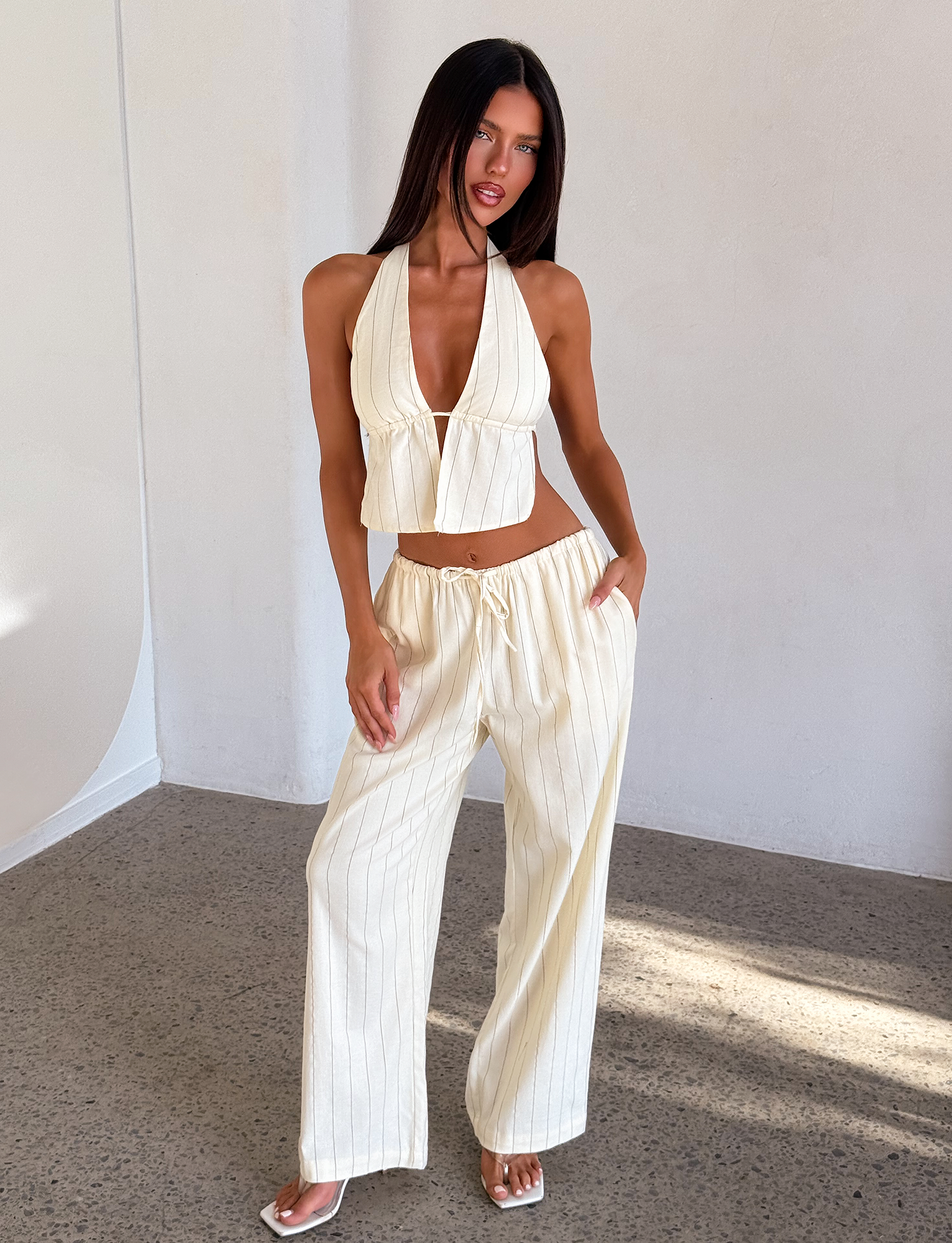 A woman poses indoors in sunlight, wearing the TIGERMIST LAINEY TOP - BEIGE STRIPE—a sleeveless, open back halter with adjustable ties—paired with matching high-waisted, wide-leg pants. One hand is in her pocket as she stands on a speckled floor.