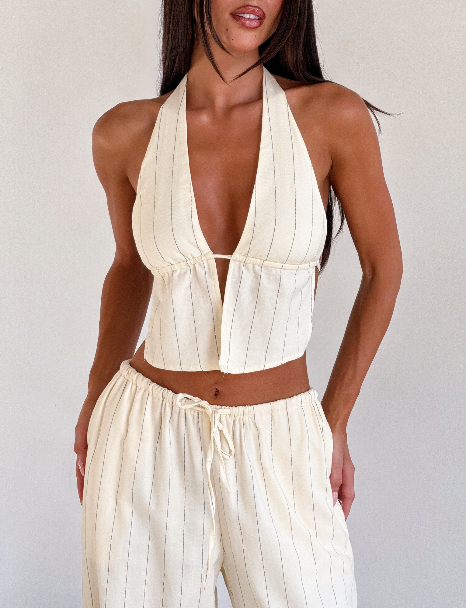 A woman wears the TIGERMIST LAINEY TOP in beige stripe with adjustable halter ties, paired with matching high-waisted, drawstring pants, standing against a plain light background.