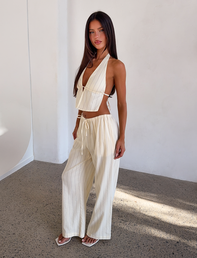 A woman with long straight hair poses indoors against a sunlit white wall, wearing the TIGERMIST LAINEY PANT - BEIGE STRIPE with wide legs, a cream backless halter top, and white sandals.