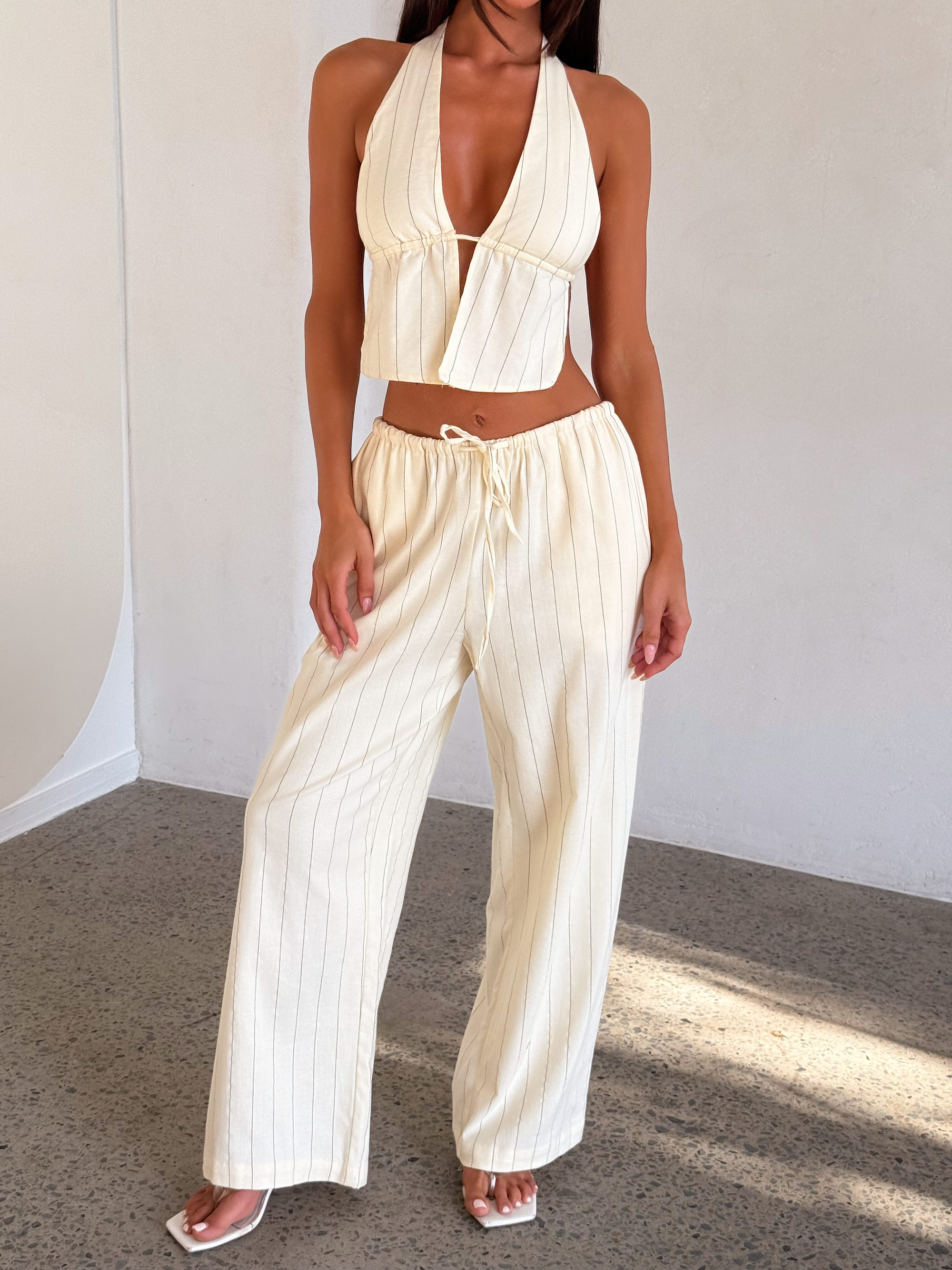 A woman, her face unseen, stands indoors on a speckled floor in natural light, wearing TIGERMIST's LAINEY PANT - BEIGE STRIPE and a matching beige pinstripe halter top.