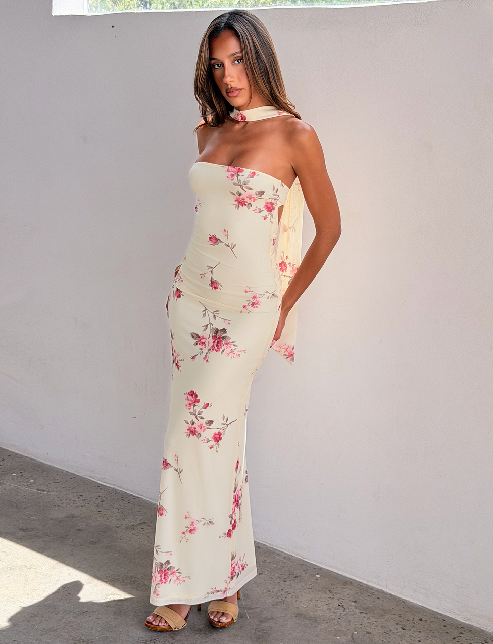 A woman wears the TIGERMIST DANIELA SCARF DRESS in yellow floral, featuring a strapless design and twisted back detail. Paired with sandals and matching scarf, she poses indoors against a white wall with one hand in her pocket.