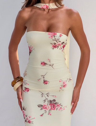 A woman wears the TIGERMIST DANIELA SCARF DRESS in yellow floral, which features a strapless fitted design with a twisted back, plus a matching choker and brown bangles, standing against a plain light background.