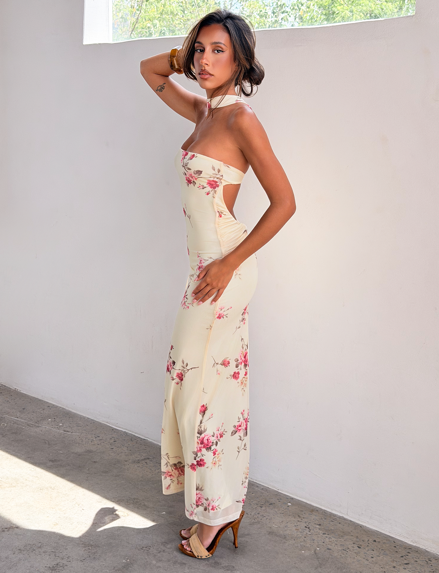 A woman models the TIGERMIST DANIELA SCARF DRESS - YELLOW FLORAL, featuring a twisted back detail, as she stands sideways in high heels against a white wall, bathed in soft sunlight from a nearby window.