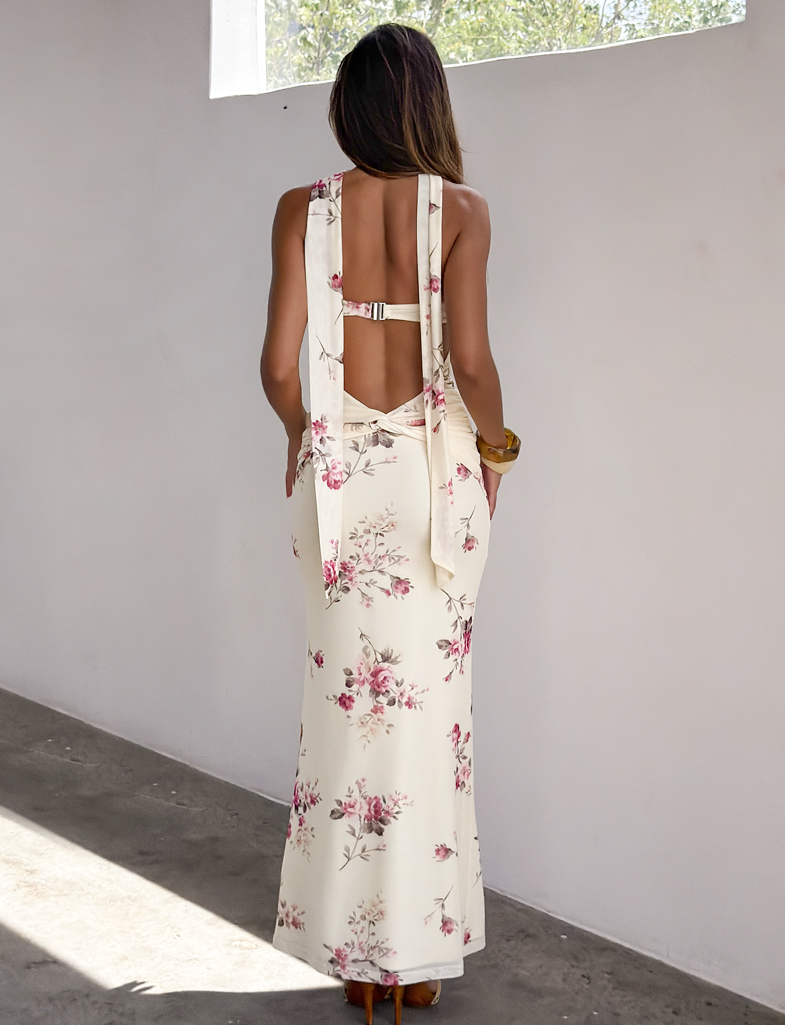 A woman stands indoors with her back to the camera, wearing the TIGERMIST DANIELA SCARF DRESS— a long, sleeveless yellow floral dress with wide straps tied into bows—while sunlight streams through a high window onto the wall.