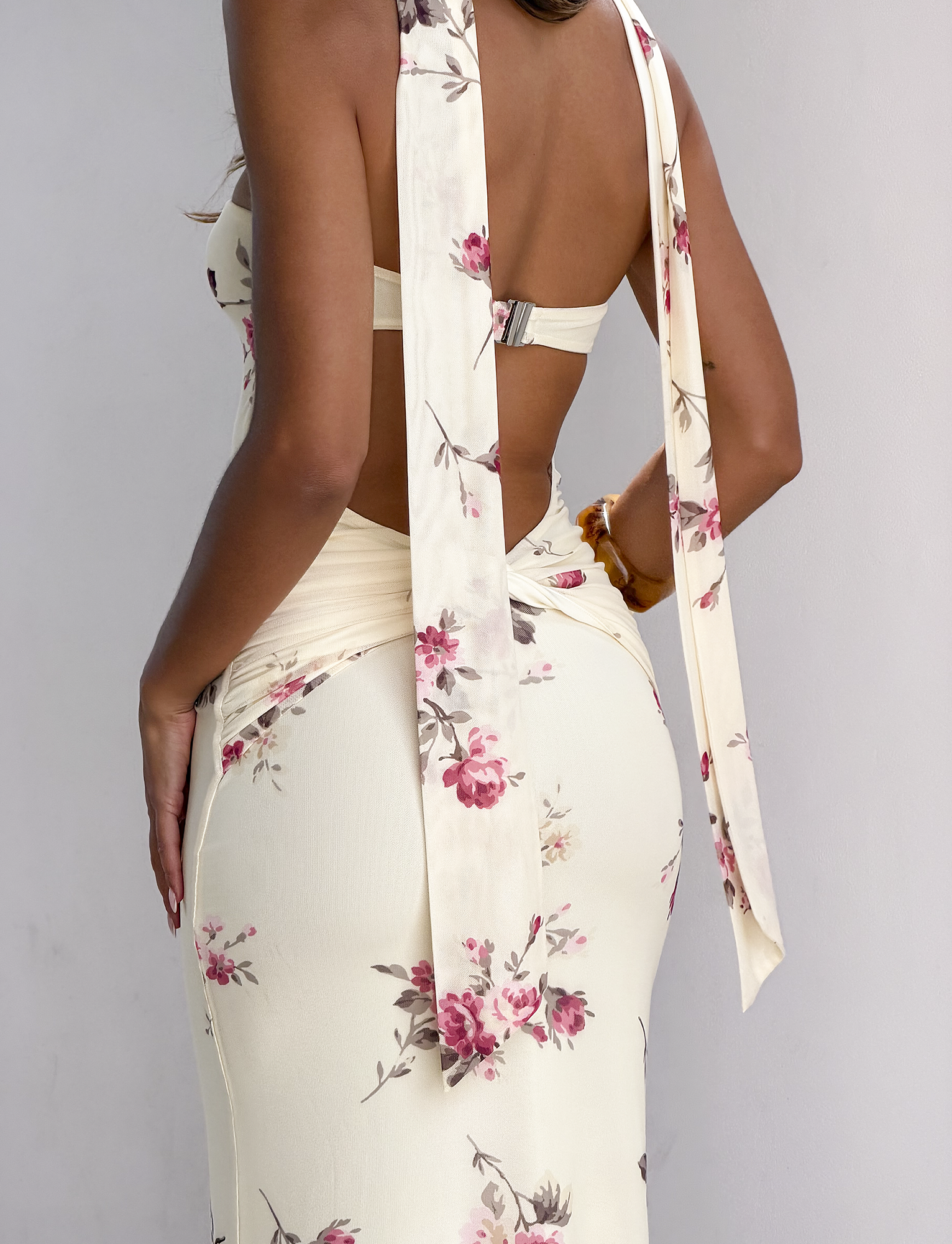 A woman stands against a white wall, shown from the back and waist down, wearing the TIGERMIST DANIELA SCARF DRESS in yellow floral, featuring long tied straps and an open back.
