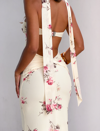 A woman stands against a white wall, shown from the back and waist down, wearing the TIGERMIST DANIELA SCARF DRESS in yellow floral, featuring long tied straps and an open back.