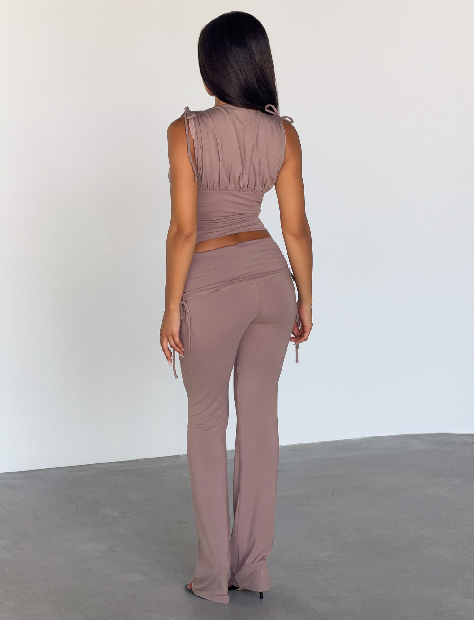 A woman with long straight hair, seen from behind indoors, wears a taupe sleeveless crop top paired with TIGERMIST's HALONA PANT in mocha, featuring a high-waisted flared silhouette against a minimal background.