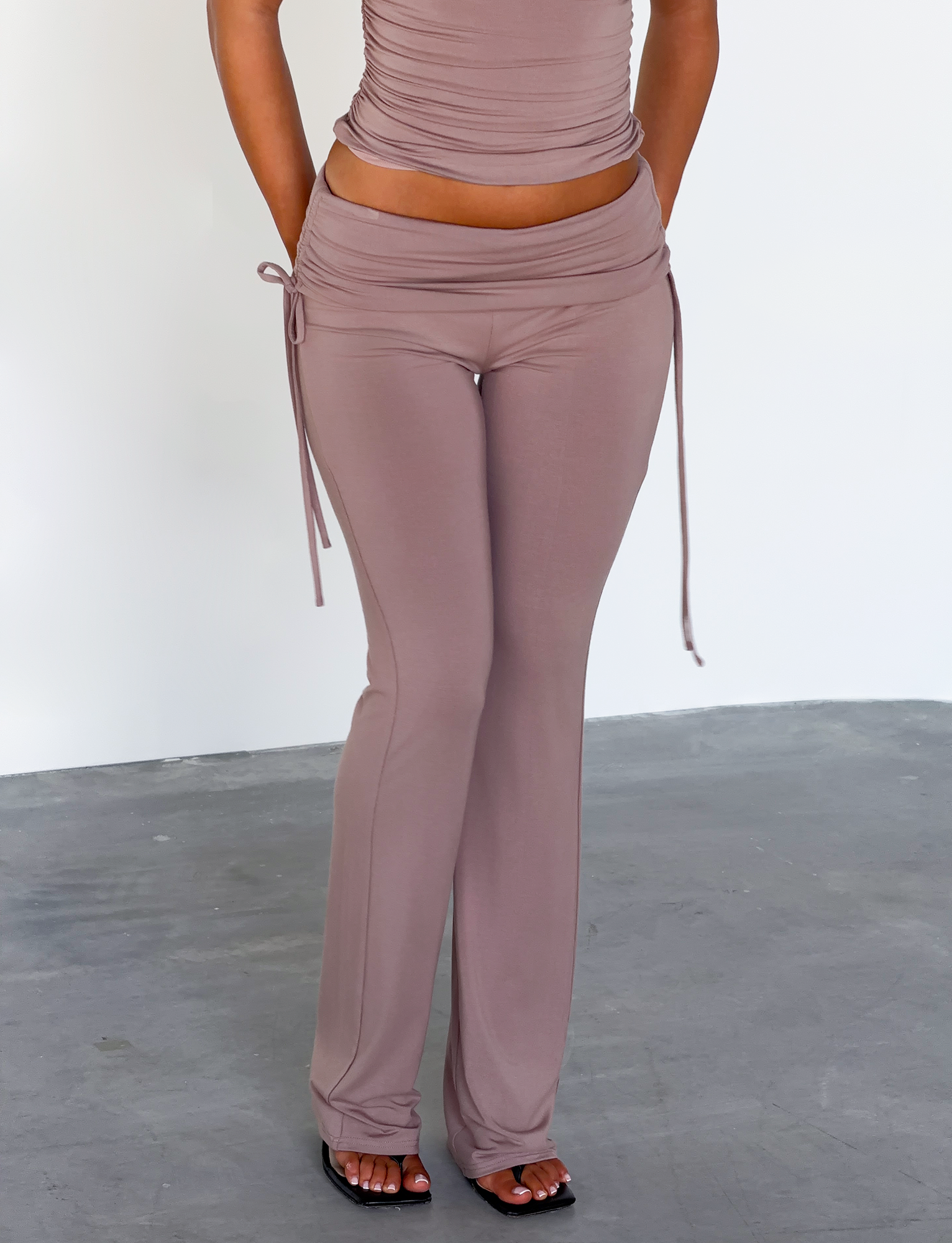 A person wears the TIGERMIST HALONA PANT in mocha, featuring a ruched waistband and flared silhouette, while standing on a gray concrete floor against a plain white wall with their head and upper torso out of frame.