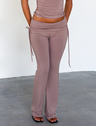 A person wears the TIGERMIST HALONA PANT in mocha, featuring a ruched waistband and flared silhouette, while standing on a gray concrete floor against a plain white wall with their head and upper torso out of frame.