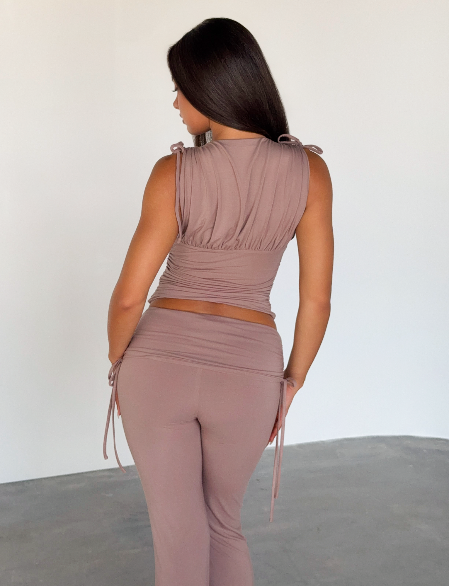 A woman with long dark hair faces away, wearing the TIGERMIST HALONA TOP in mocha—a crop top with adjustable shoulder ties—paired with matching high-waisted pants, standing on concrete before a plain white background.