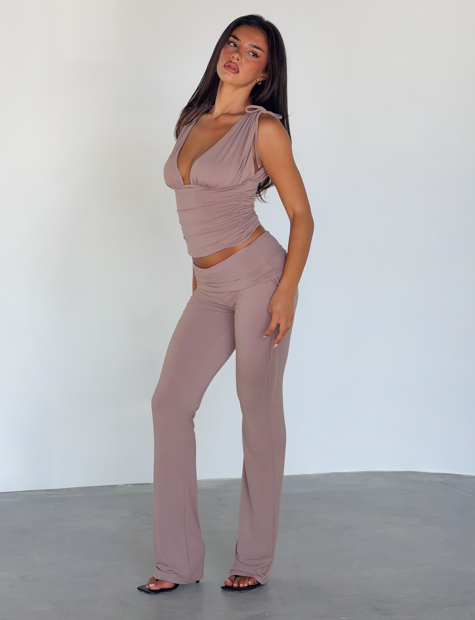 A woman with long dark hair stands confidently against a light background, wearing TIGERMIST’s HALONA PANT in mocha, styled with a mauve sleeveless deep-neck top and black heeled sandals.