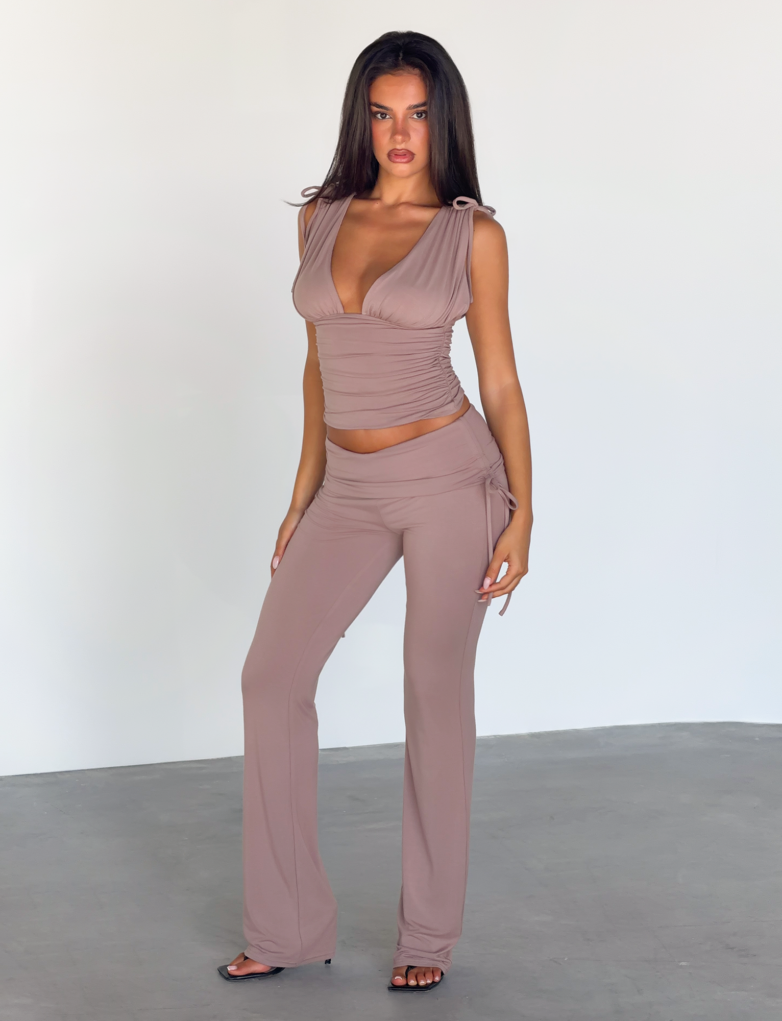 A woman with long dark hair poses indoors, wearing the TIGERMIST HALONA TOP in mocha with a ruched bodice and plunging neckline, paired with matching high-waisted, flared pants. She stands on a gray floor against a plain white background.