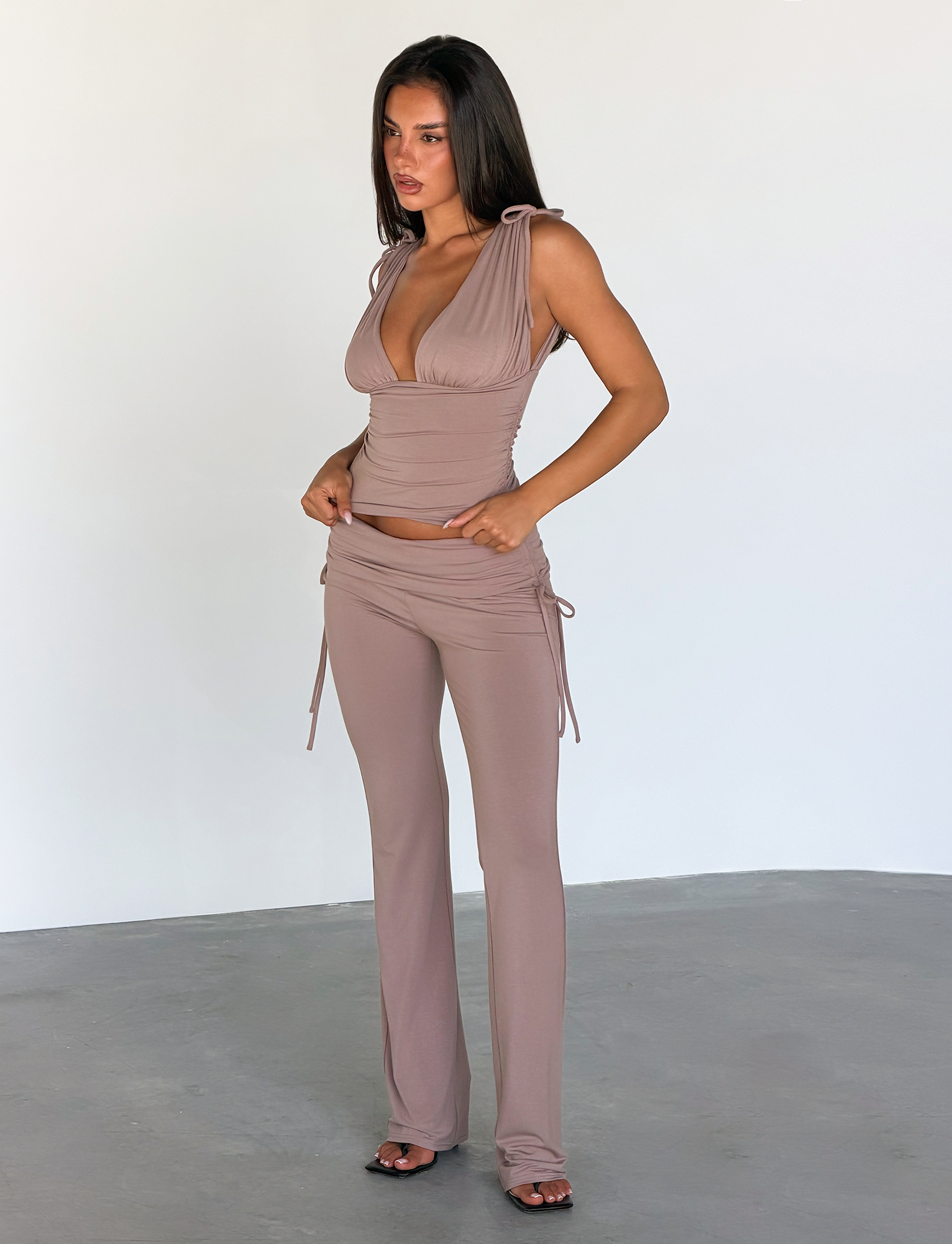 A woman stands indoors wearing the TIGERMIST HALONA PANT in mocha, featuring a ruched waistband and flared silhouette, styled with a taupe sleeveless deep v-neck top and black open-toe shoes against a plain light background.