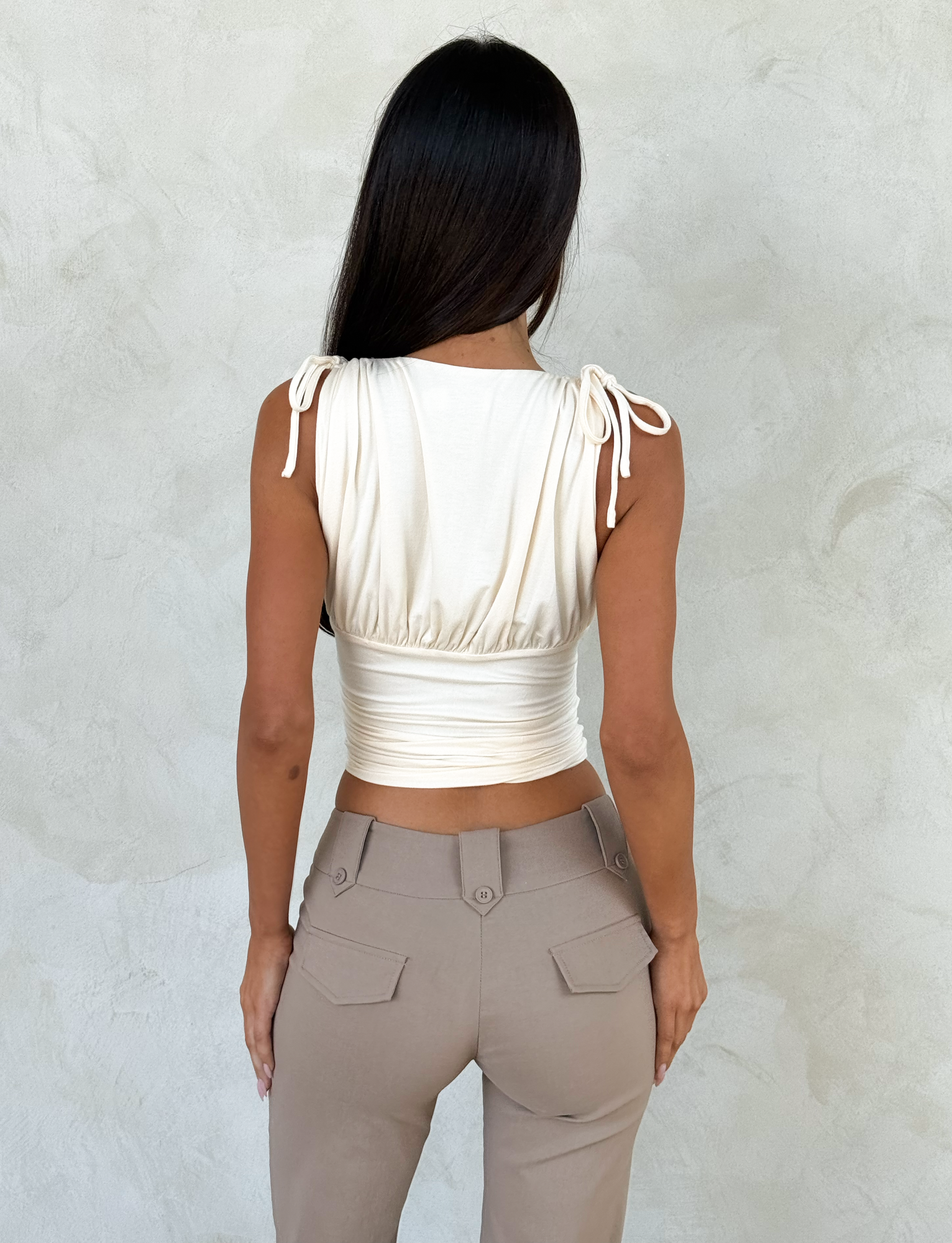 A woman with long dark hair, seen from behind, wears the TIGERMIST HALONA TOP in cream with adjustable shoulder ties and high-waisted taupe pants, standing against a light textured wall.