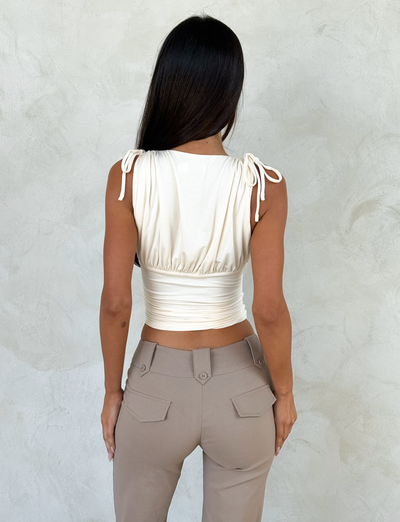 A woman with long dark hair, seen from behind, wears the TIGERMIST HALONA TOP in cream with adjustable shoulder ties and high-waisted taupe pants, standing against a light textured wall.